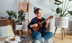Młoda kobieta gra na ukulele, siedząc na podłodze. Czy gra na ukulele jest trudna? Ona wygląda na zrelaksowaną i szczęśliwą.
