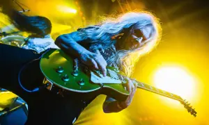 Długowłosy gitarzysta gra na zielonej gitarze, a w tle widać światła sceniczne. Tekst "turbo dorosłe dzieci" idealnie pasuje do tej rockowej sceny.
