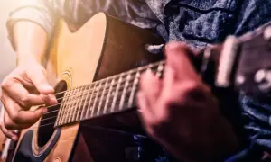 Ręce grające na gitarze akustycznej. Zbliżenie na palce naciskające struny i kostkę. Idealny moment, by zacząć naukę, jak nauczyć się grać na gitarze.
