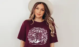 Kobieta w kapeluszu nosi koszulkę z napisem "Take Me Home Country Roads" i grafiką kowbojskich butów.