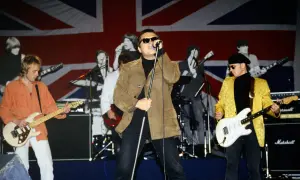 Zespół gra na scenie z flagą Wielkiej Brytanii w tle. Wokalista w brązowej kurtce, gitarzyści w pomarańczowej i żółtej. king t love.
