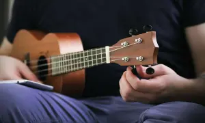 Ręka kręci kluczem, by nastroić ukulele sopranowe. Na kolanach leży tablet z instrukcją.