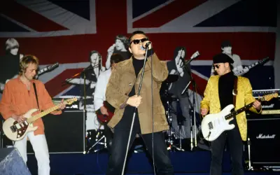 Zespół gra na scenie z flagą Wielkiej Brytanii w tle. Wokalista w brązowej kurtce, gitarzyści w pomarańczowej i żółtej. king t love.