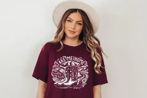 Kobieta w kapeluszu nosi koszulkę z napisem "Take Me Home Country Roads" i grafiką kowbojskich butów.