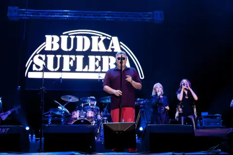 Budka Suflera na scenie. Wokalista w okularach śpiewa, a obok niego dwie wokalistki. W tle perkusja i logo zespołu.