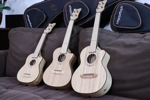 Trzy ukulele sopranowe z jasnego drewna leżą na brązowej kanapie obok czarnych futerałów.