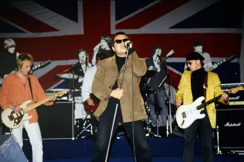 Zespół gra na scenie z flagą Wielkiej Brytanii w tle. Wokalista w brązowej kurtce, gitarzyści w pomarańczowej i żółtej. king t love.