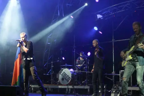 Zespół Lemon na scenie, wokalista z flagą, gitarzysta basowy, perkusja z logo.