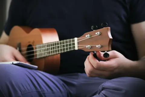 Ręka kręci kluczem, by nastroić ukulele sopranowe. Na kolanach leży tablet z instrukcją.