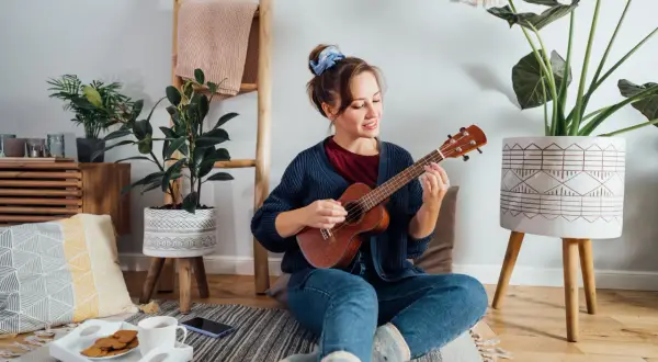 Młoda kobieta gra na ukulele, siedząc na podłodze. Czy gra na ukulele jest trudna? Ona wygląda na zrelaksowaną i szczęśliwą.