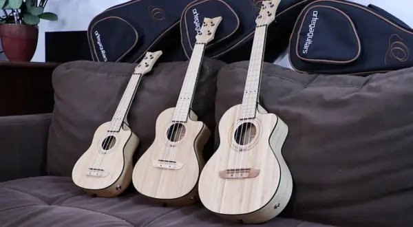 Trzy ukulele sopranowe z jasnego drewna leżą na brązowej kanapie obok czarnych futerałów.