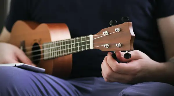 Ręka kręci kluczem, by nastroić ukulele sopranowe. Na kolanach leży tablet z instrukcją.