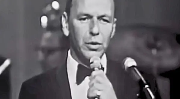 Frank Sinatra śpiewa "Fly Me to the Moon lyrics", ubrany w smoking.