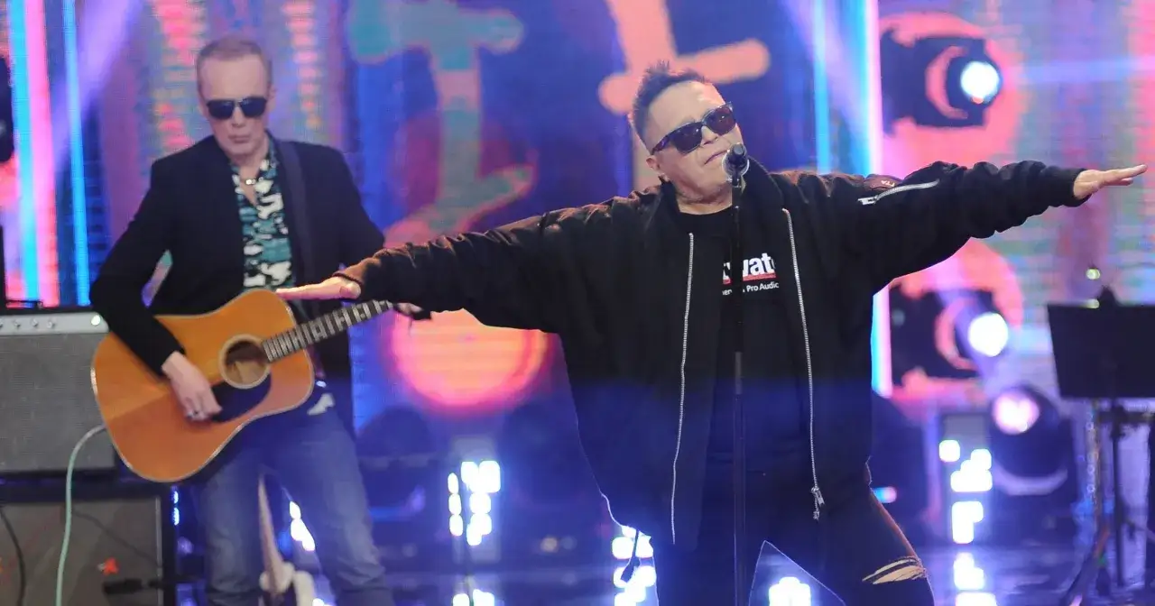 King T-Love na scenie, w czarnej kurtce, z rozłożonymi ramionami. Obok niego gitarzysta.