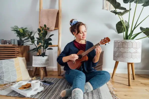 Młoda kobieta gra na ukulele, siedząc na podłodze. Czy gra na ukulele jest trudna? Ona wygląda na zrelaksowaną i szczęśliwą.