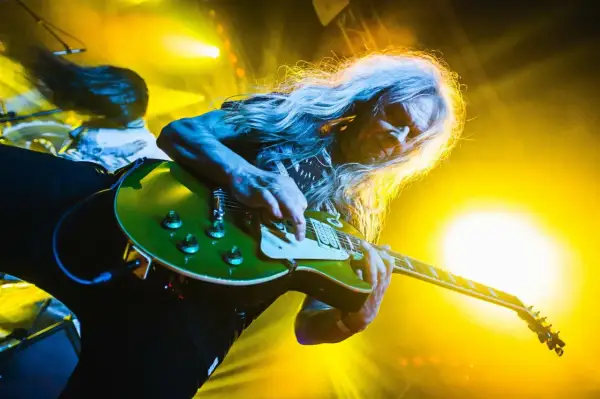 Długowłosy gitarzysta gra na zielonej gitarze, a w tle widać światła sceniczne. Tekst "turbo dorosłe dzieci" idealnie pasuje do tej rockowej sceny.
