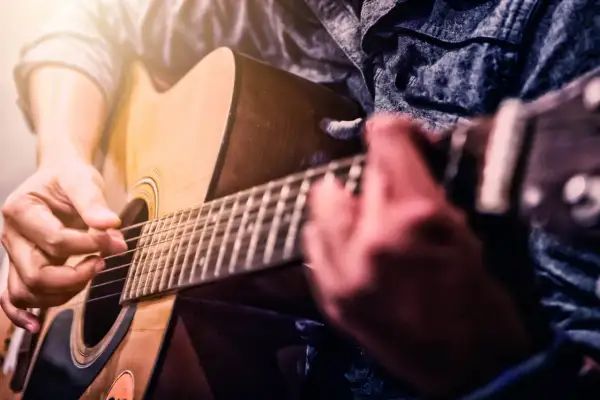 Ręce grające na gitarze akustycznej. Zbliżenie na palce naciskające struny i kostkę. Idealny moment, by zacząć naukę, jak nauczyć się grać na gitarze.