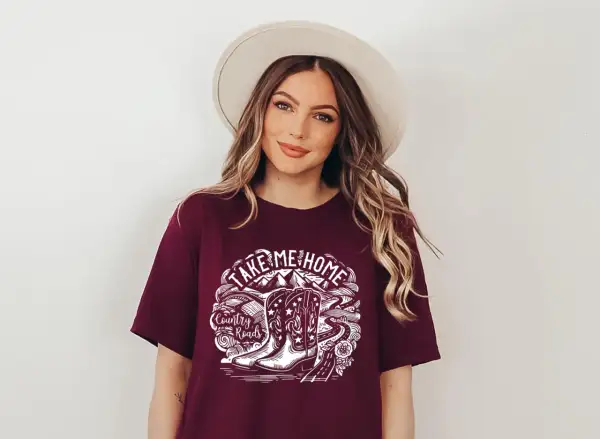 Kobieta w kapeluszu nosi koszulkę z napisem "Take Me Home Country Roads" i grafiką kowbojskich butów.