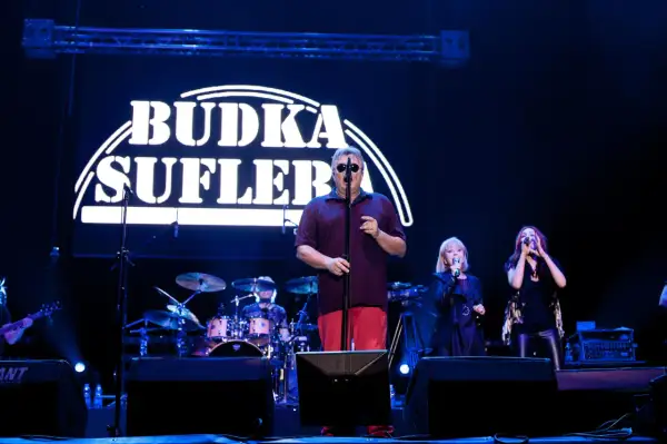 Budka Suflera na scenie. Wokalista w okularach śpiewa, a obok niego dwie wokalistki. W tle perkusja i logo zespołu.