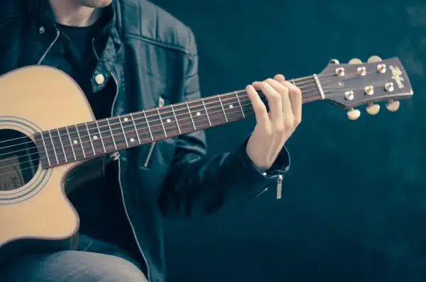 Mężczyzna w skórzanej kurtce uczy się, jak szarpać struny na gitarze. Jego palce zaciskają akord na gryfie.