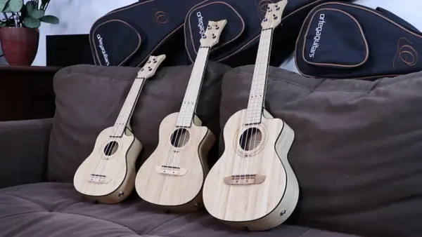 Trzy ukulele sopranowe z jasnego drewna leżą na brązowej kanapie obok czarnych futerałów.